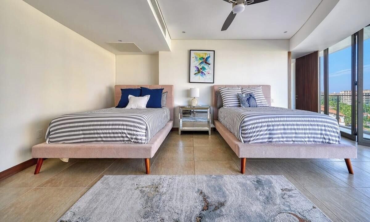 A modern bedroom with two beds, a ceiling fan, a large window, and a city view.