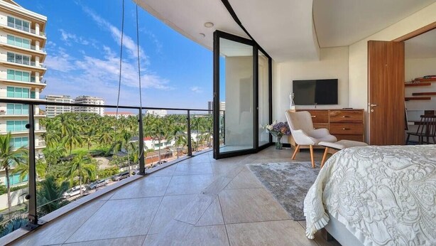 Un dormitorio con una cama grande, televisión y balcón con vista a palmeras y edificios.