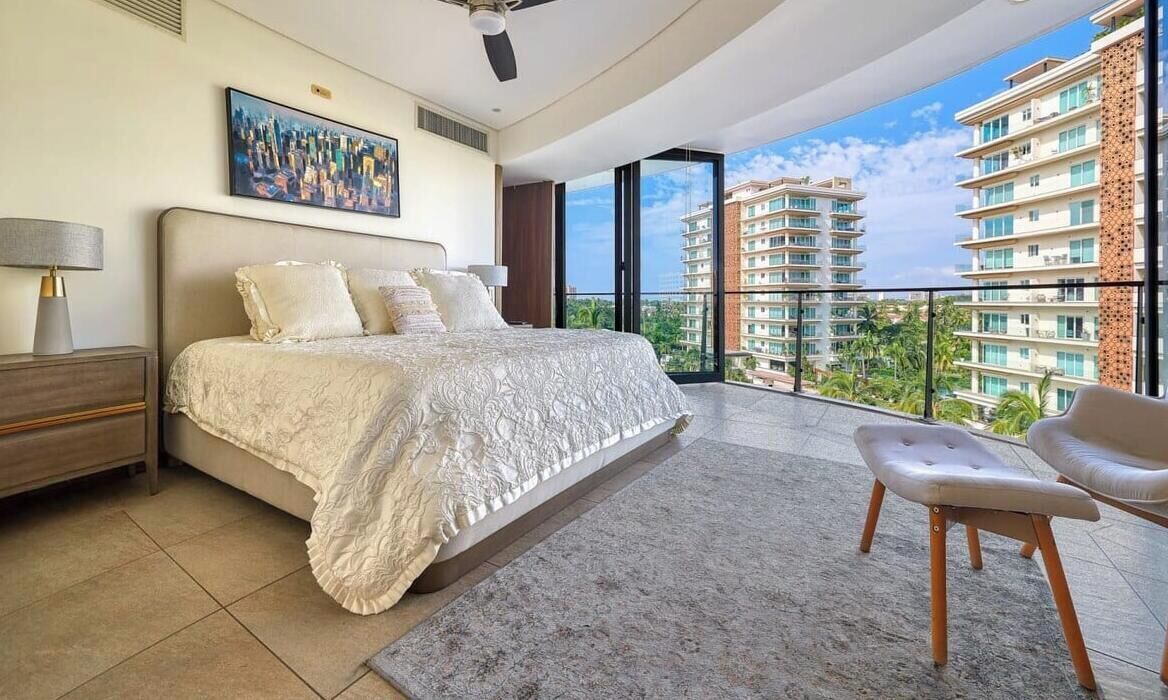 Un dormitorio con una cama grande, una mesita de noche, una silla y un balcón con vistas a la ciudad.