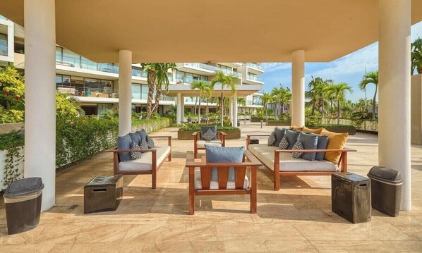 A covered outdoor seating area with wooden furniture and cushions, surrounded by palm trees and a modern building in the background. - Luxury 2BR Condo in Marina - Pool - Zen Rooftop (Puerto Vallarta)