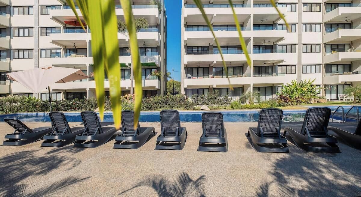 Uma área na beira da piscina com espreguiçadeiras e um edifício ao fundo.