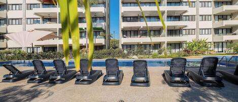 Uma área na beira da piscina com espreguiçadeiras e um edifício ao fundo.