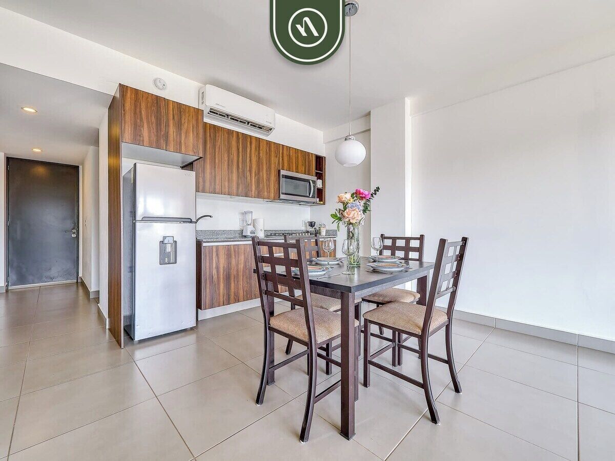 A modern kitchen with wooden cabinets, a built-in microwave, and a refrigerator. There is a dining table with four chairs and a vase with flowers.