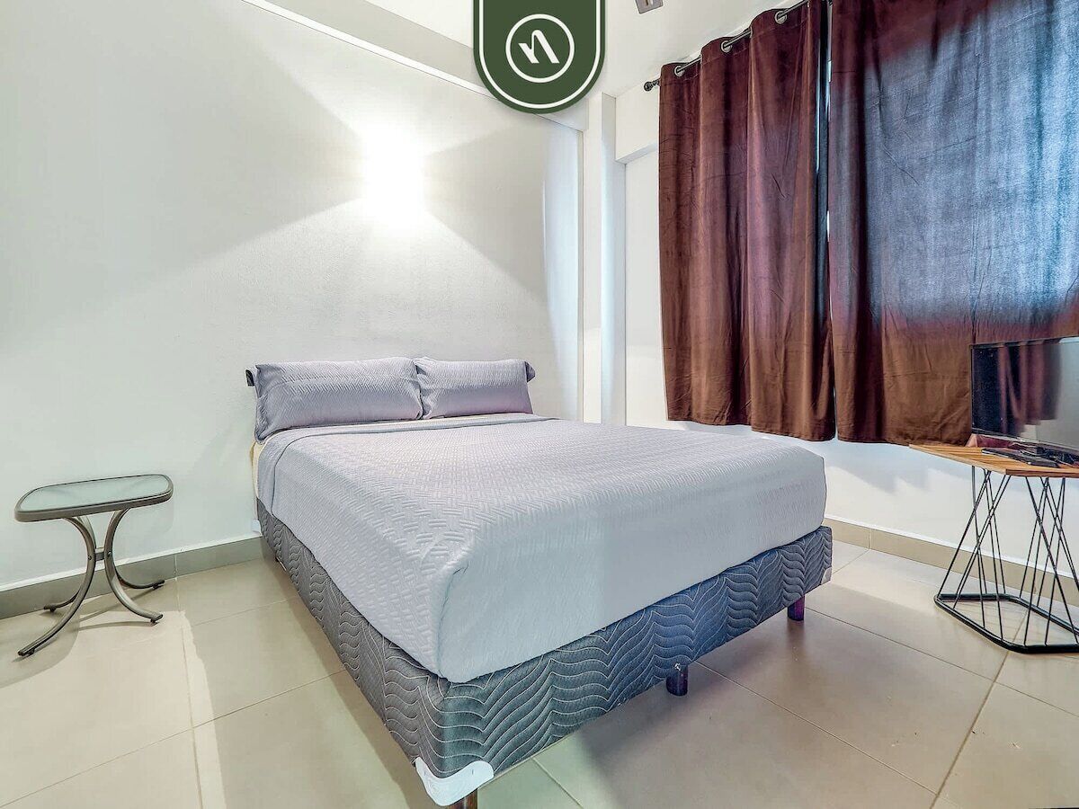 A bedroom with a bed, a small table, a TV, and a window with curtains.