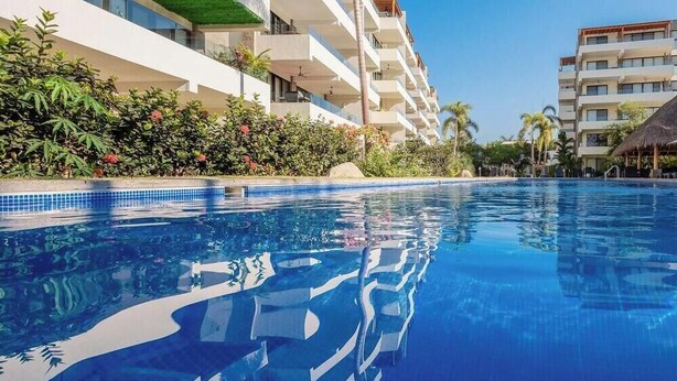 A swimming pool with clear blue water, surrounded by hotel buildings and lush greenery.