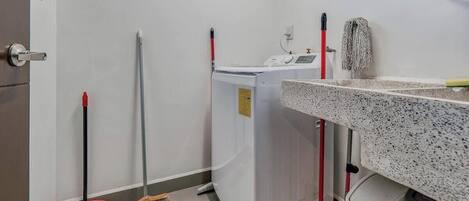 A laundry room with a washing machine, a mop, a broom, and a trash bin.