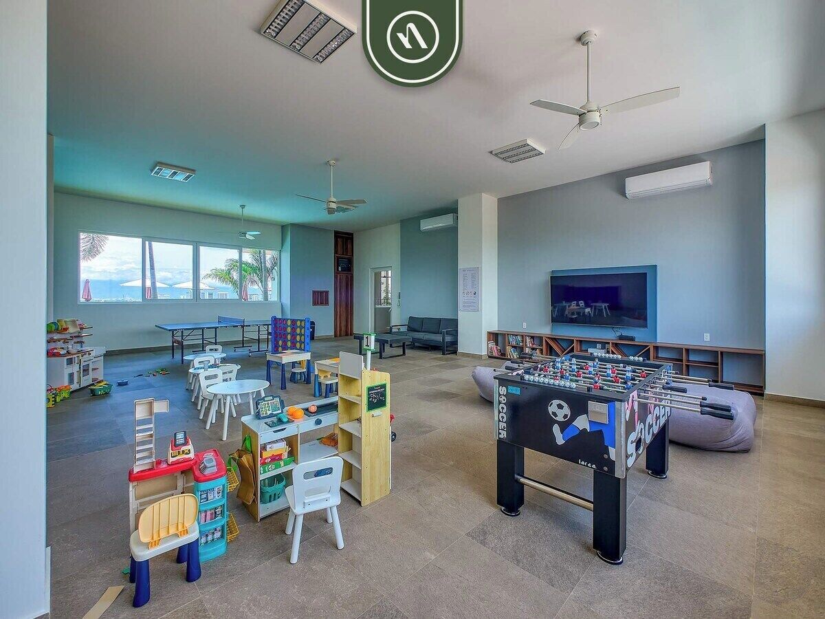 A spacious room with a foosball table, tables with chairs, and a large window.