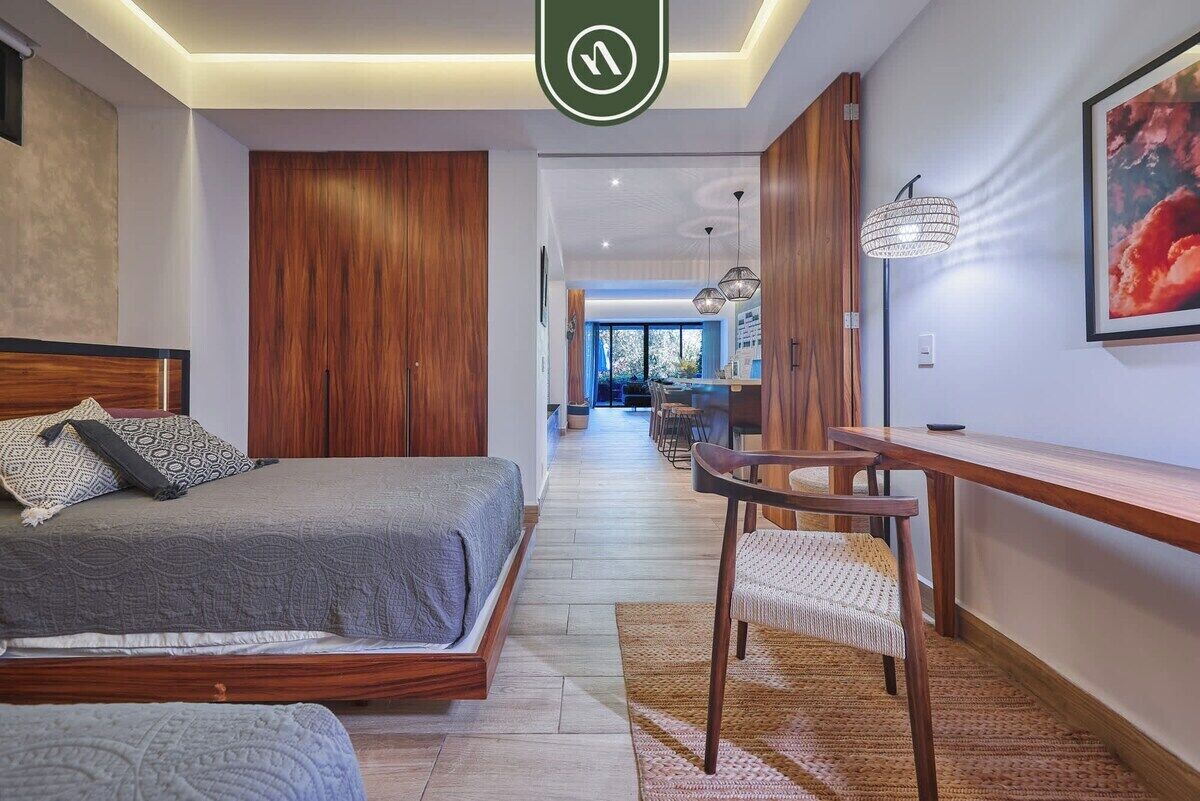 A modern bedroom with a bed, a desk, and a chair. There is a hallway leading to a dining area.