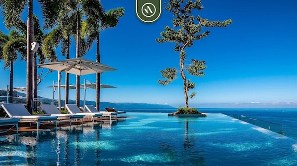 Une piscine à débordement bordée de chaises longues et d’ombrelles, entourée de palmiers et offrant une vue sur l’océan.