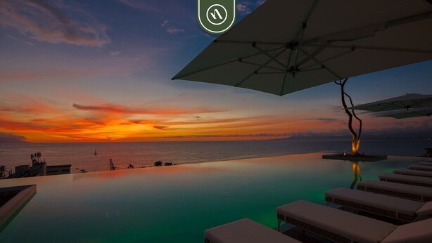 Ein Infinity-Pool mit Liegestühlen und Sonnenschirmen mit Blick auf einen Sonnenuntergang über dem Meer.