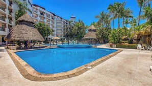 A swimming pool surrounded by palm trees and thatched-roof structures, with a multi-story hotel building in the background. - 3BR Condo With Private Balcony - Pool - Marina` (Puerto Vallarta)