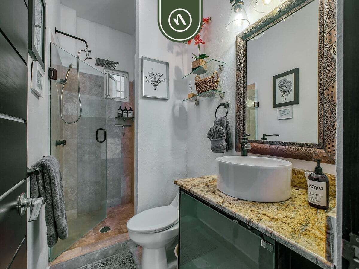 A modern bathroom with a glass shower enclosure, a white toilet, a large sink with a marble countertop, and a mirror with a decorative frame.
