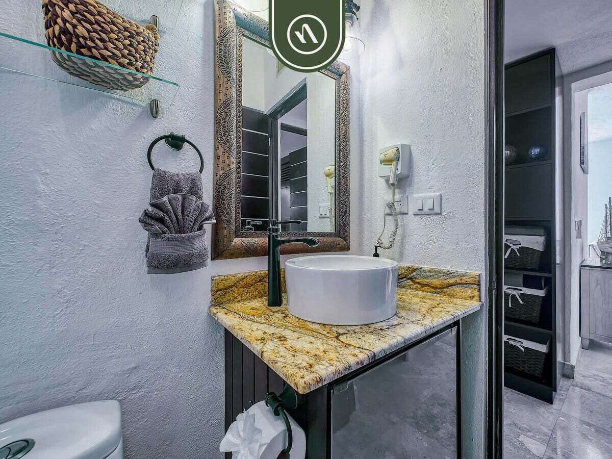 Un baño con encimera de mármol, lavamanos blanco, aro para toallas, espejo y una cesta en la pared.