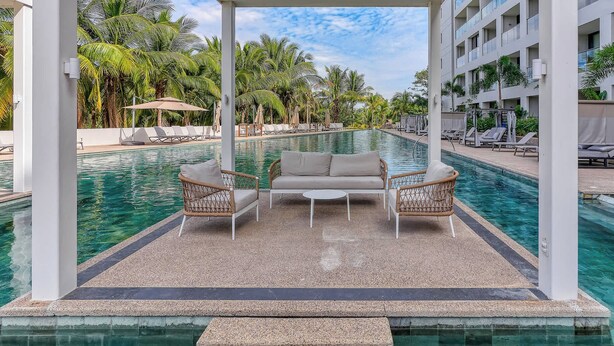 Zona de piscina con área de descanso cubierta, sillones y vista a la piscina y palmeras circundantes.