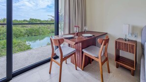 Un comedor con una mesa preparada para dos personas, dos sillas, una mesita auxiliar con una lámpara, y vistas a la vegetación y al agua a través de grandes ventanales.