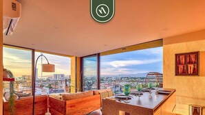 A modern interior with a sofa, dining table, and a view of the cityscape. - Brand New 2BR APT - Stunning Ocean & Sunset Views (Puerto Vallarta)