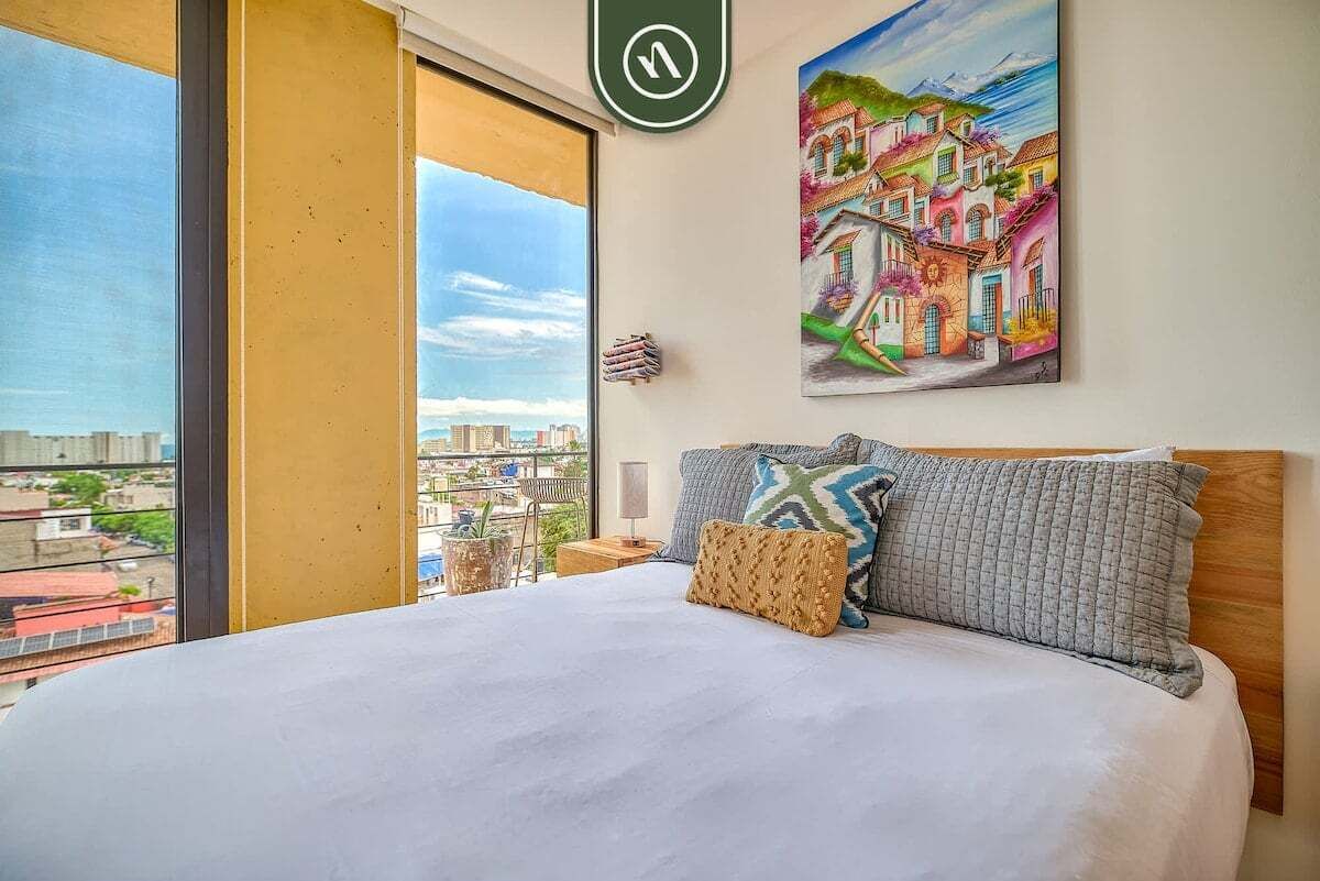 Habitació d'hotel amb un llit gran, un quadre de construccions de colors a la paret i una vista de la ciutat a través d'una finestra gran.