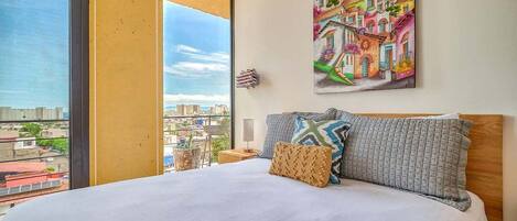 Habitació d'hotel amb un llit gran, un quadre de construccions de colors a la paret i una vista de la ciutat a través d'una finestra gran.