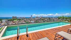A swimming pool with a wooden deck, lounge chairs, and a cityscape view. - Brand New 2BR APT - Stunning Ocean & Sunset Views (Puerto Vallarta)