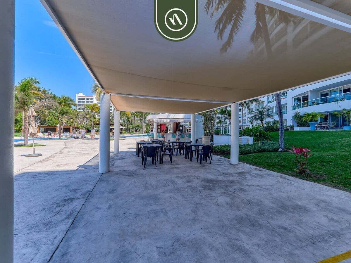 An outdoor seating area with tables and chairs under a shaded canopy, surrounded by greenery and a building in the background.