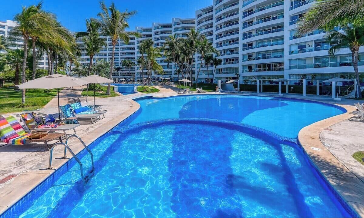 A swimming pool with lounge chairs and umbrellas, surrounded by palm trees and a multi-story building.