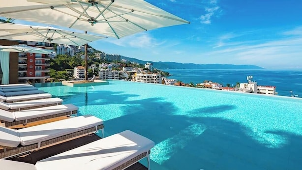 Una piscina infinita azul cristalina con sillones de descanso y sombrillas, con vistas a una ciudad costera.