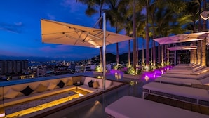 Un área de lounge en la azotea con asientos y sombrillas, con vistas a un paisaje urbano al atardecer.