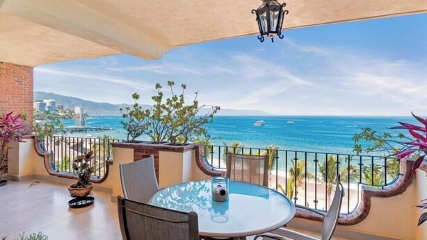Ein Balkon mit Glastisch und Stühlen, der Blick auf die Küste mit Booten und einem klaren Himmel bietet.