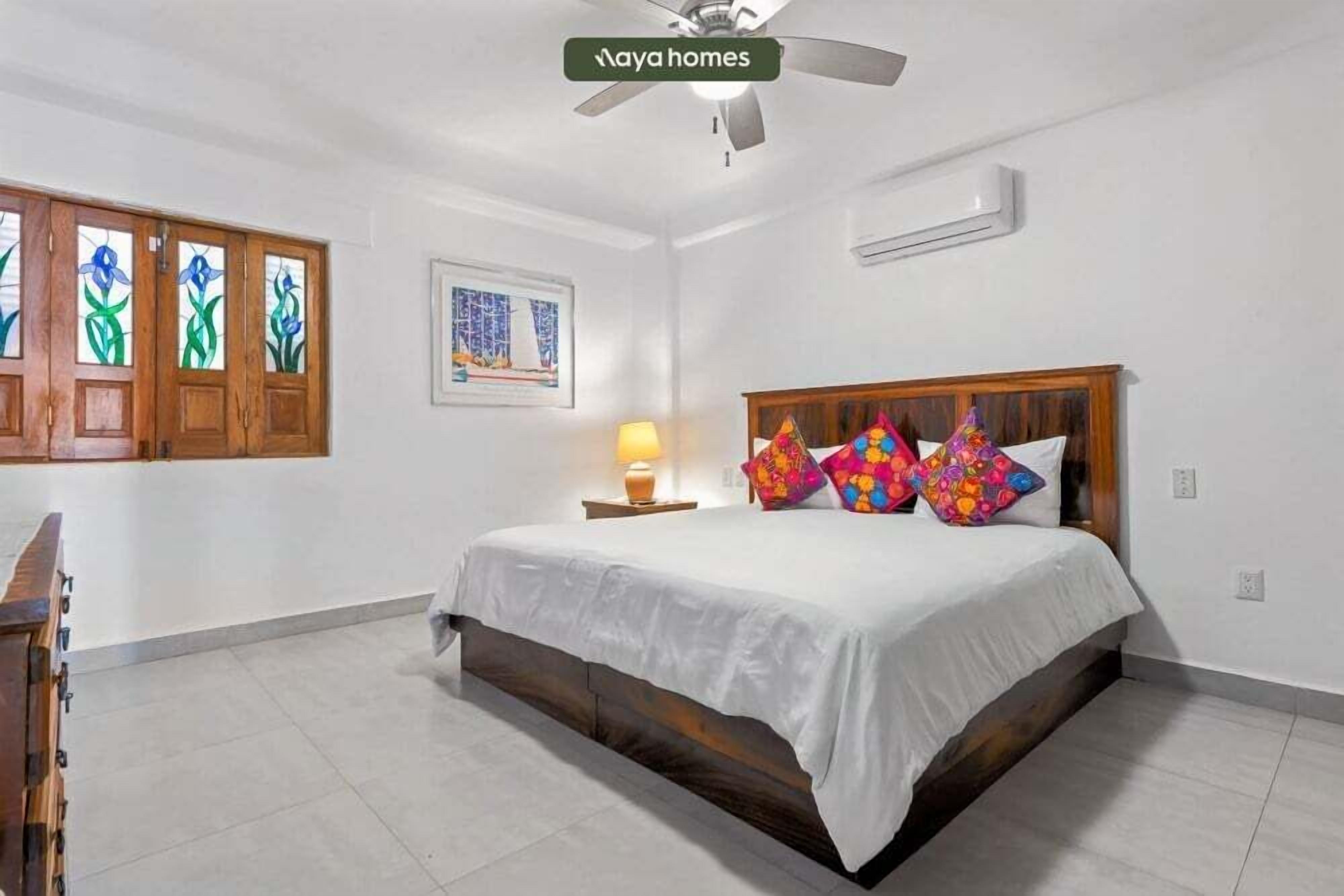 A bedroom with a bed, a ceiling fan, a nightstand with a lamp, and a window with stained glass.