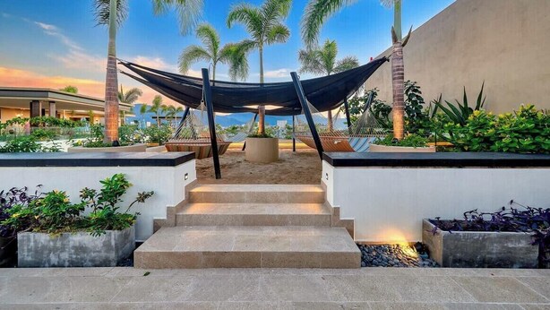 A modern outdoor area with a staircase leading to a seating area under a canopy, surrounded by palm trees and plants.