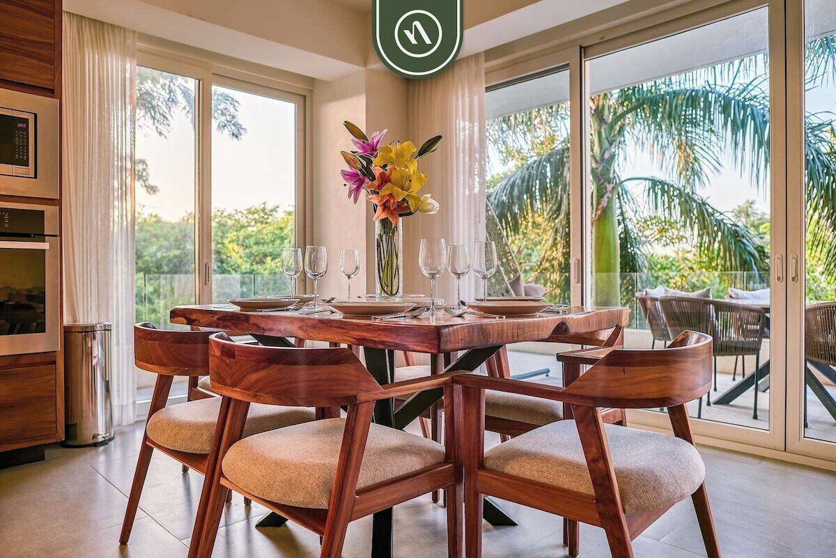 A dining area with a wooden table and chairs, a vase with flowers, and a view of palm trees through large windows.