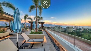 A rooftop terrace with lounge chairs, umbrellas, and a view of the cityscape and mountains.