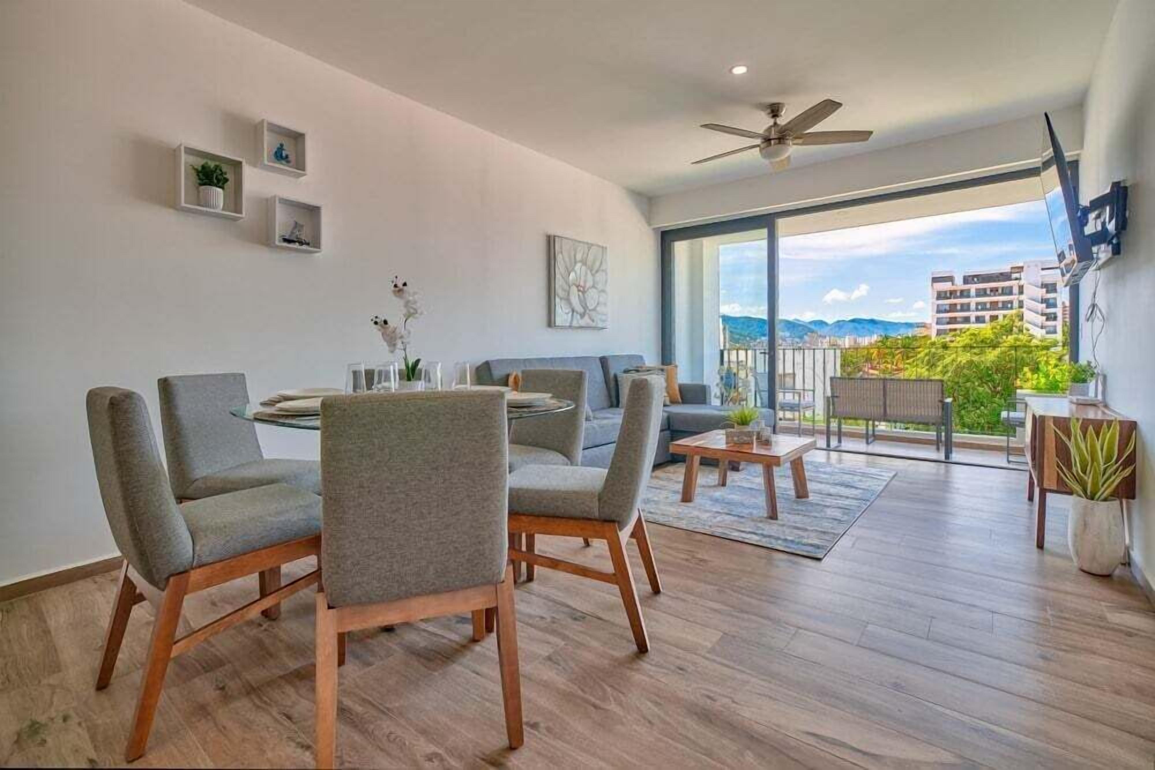 A modern living room with a dining area, a ceiling fan, and a balcony with a city view.