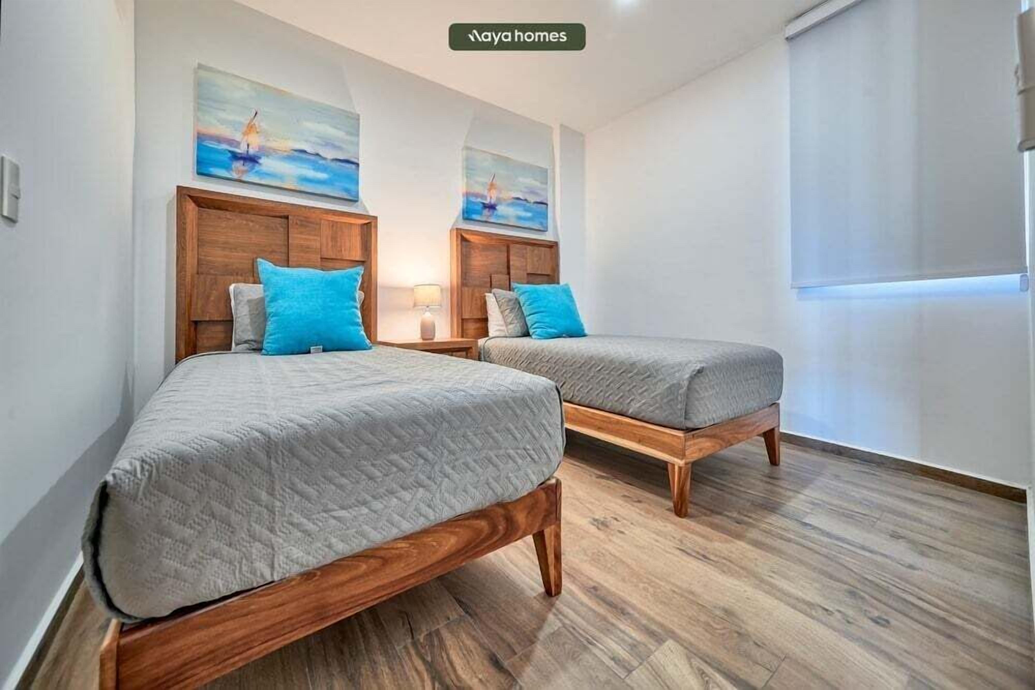 Ein Zimmer mit zwei Holzbetten, jeweils mit grauer Matratze und blauen Kissen, sowie einem Holz-Kopfteil. Darüber an der Wand befinden sich zwei Gemälde, und rechts ein Fenster mit weißem Rollo.