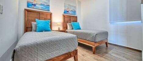 Ein Zimmer mit zwei Holzbetten, jeweils mit grauer Matratze und blauen Kissen, sowie einem Holz-Kopfteil. Darüber an der Wand befinden sich zwei Gemälde, und rechts ein Fenster mit weißem Rollo.