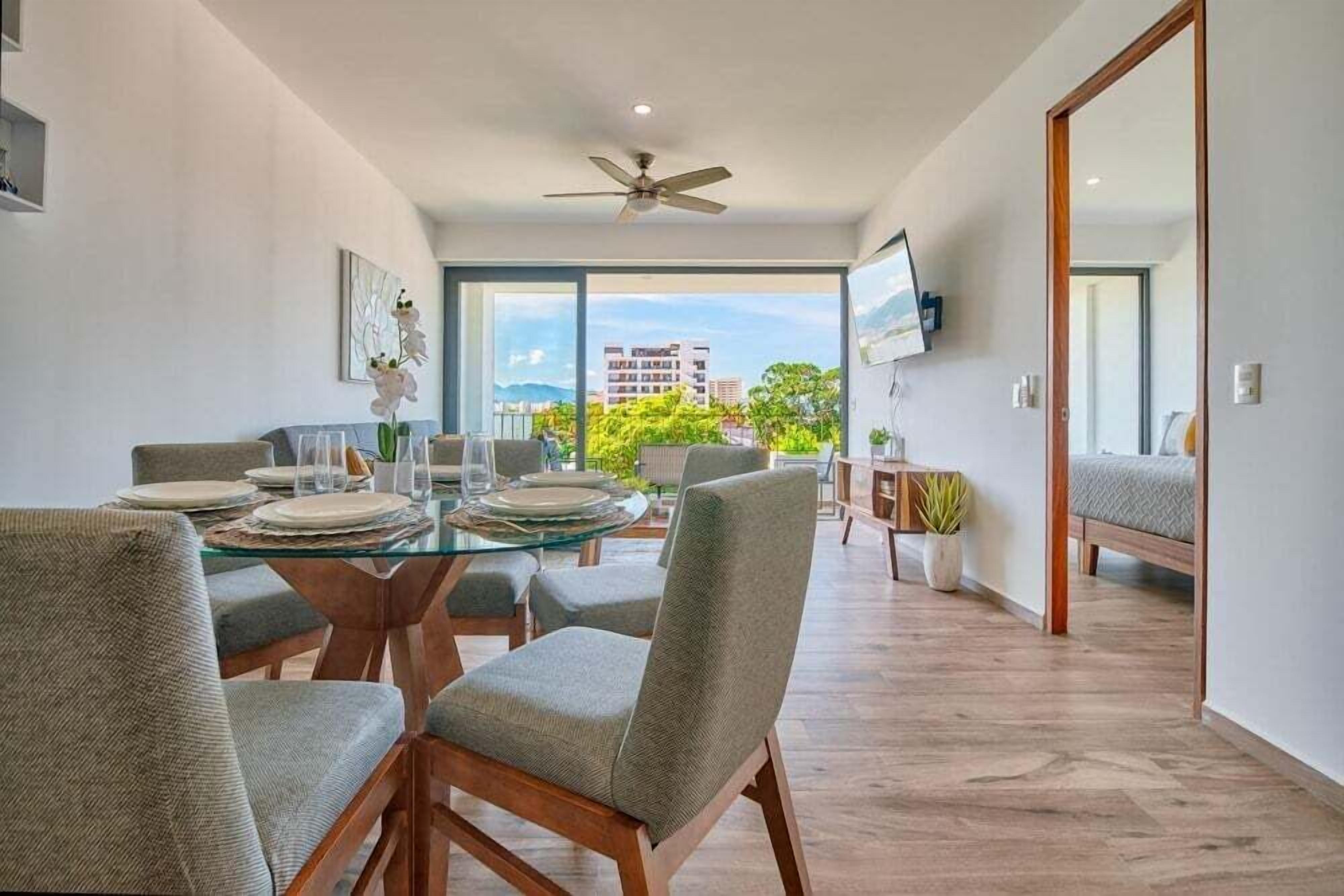 A modern dining area with a glass table, grey upholstered chairs, and a ceiling fan.