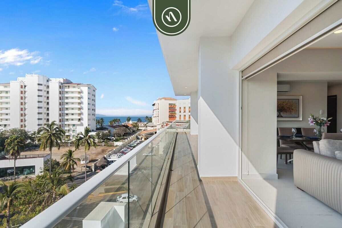 A balcony with a view of a beach and buildings, a dining area with a table and chairs, and a modern interior design.
