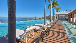 A poolside area with lounge chairs, palm trees, and a view of the sea and distant buildings. - Two Story 2 BR Penthouse - Pool - Ocean View (Puerto Vallarta)