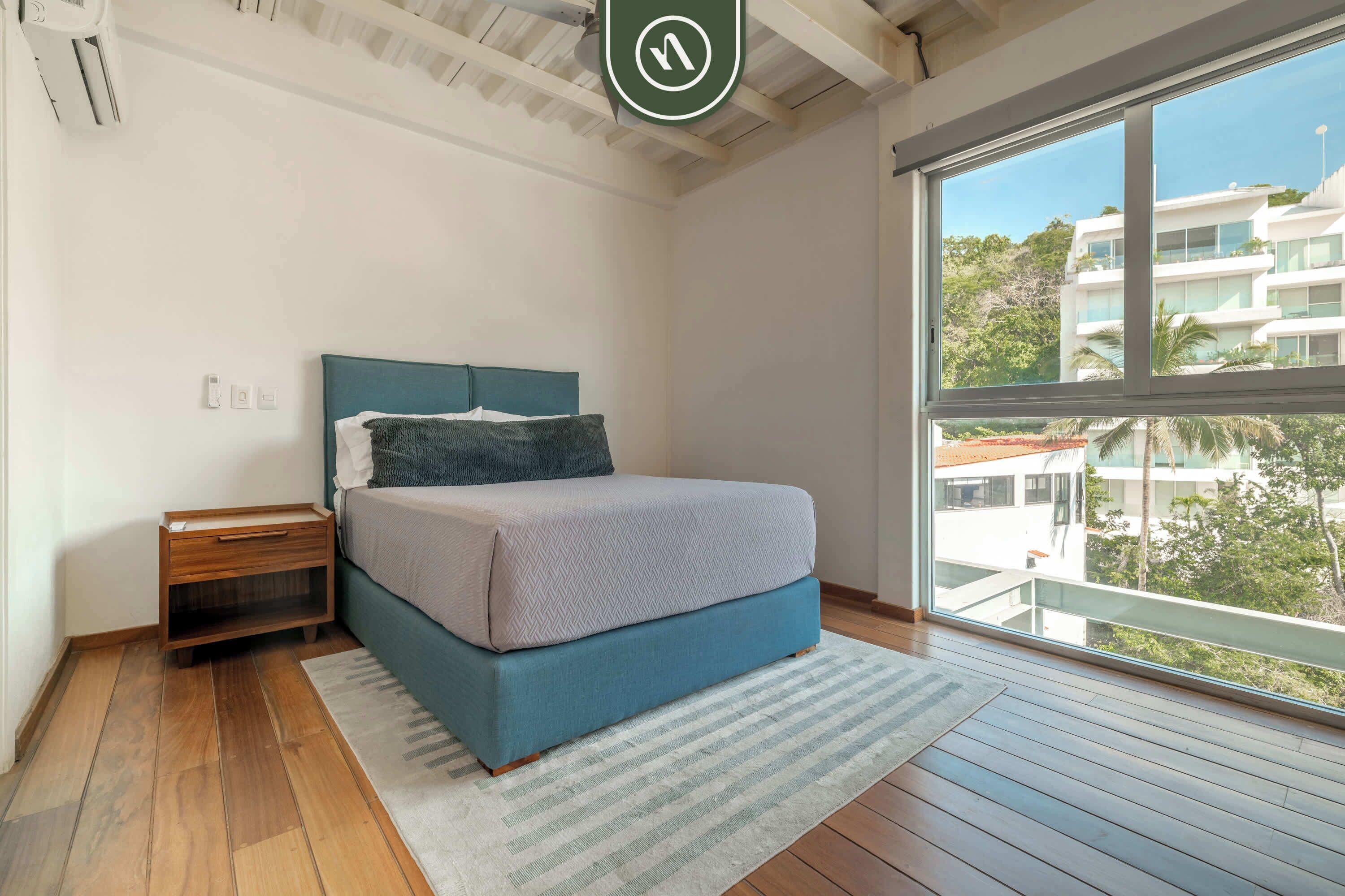 A bedroom with a bed, nightstand, and a large window with a view of buildings.