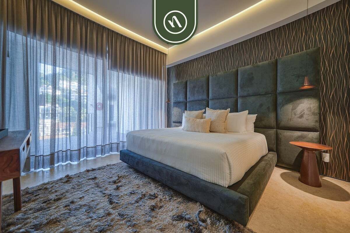 A modern bedroom with a large bed, a headboard, a small table, and a view of the outdoors through sheer curtains.