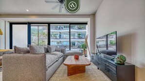 A modern living room with a sofa, coffee table, and TV, large windows with a view of a building. - Luxury Studio in Romantic Zone - Infinity Pool (Puerto Vallarta)