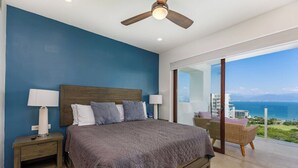 Un dormitorio con una cama grande, dos mesitas de noche con lámparas, un ventilador de techo y vista al océano a través de un ventanal corredero.