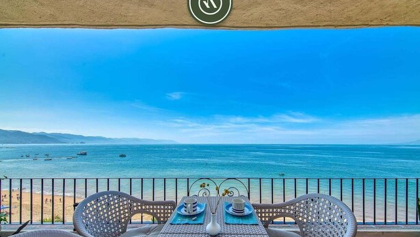 A balcony with wicker furniture, a table set for two, and a view of the sea and beach. - Brand New 2BR Condo With Oceanview - Terrace (Puerto Vallarta)