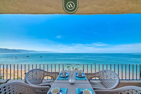 A balcony with wicker furniture, a table set with a vase and cups, and a view of the sea and beach.