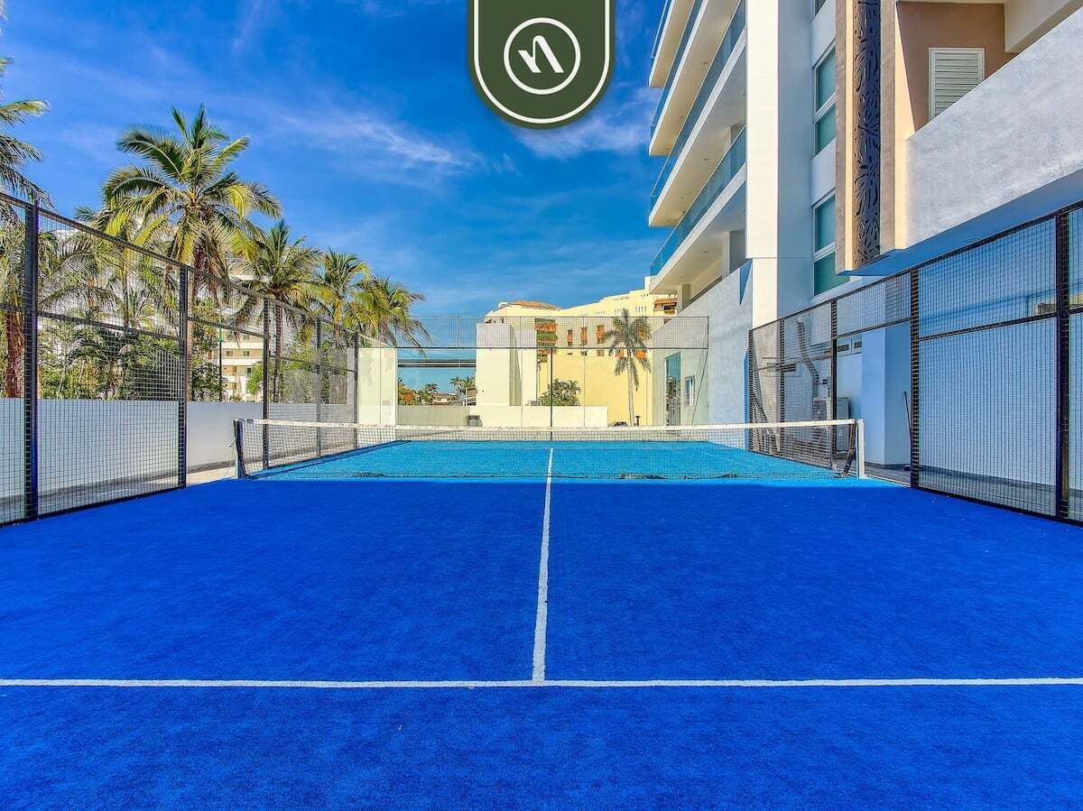 A blue tennis court with a net, surrounded by a fence, palm trees, and buildings in the background.