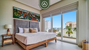 A bedroom with a large bed, a nightstand, a lamp, a painting, and a view of buildings and palm trees through large windows.
