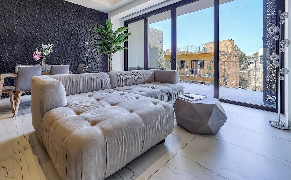 A modern living room with a sectional sofa, a dining table with chairs, a large window with a view of buildings, and a decorative wall.