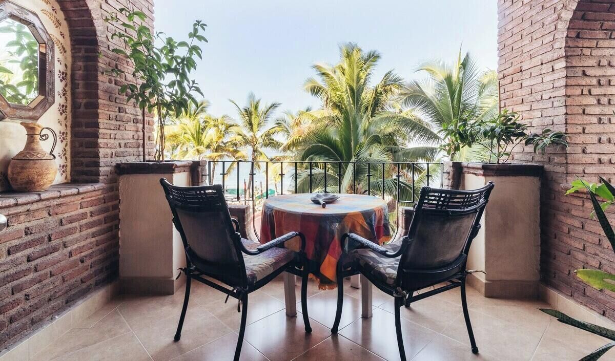 A balcony with two chairs and a table, brick walls, and a view of palm trees.