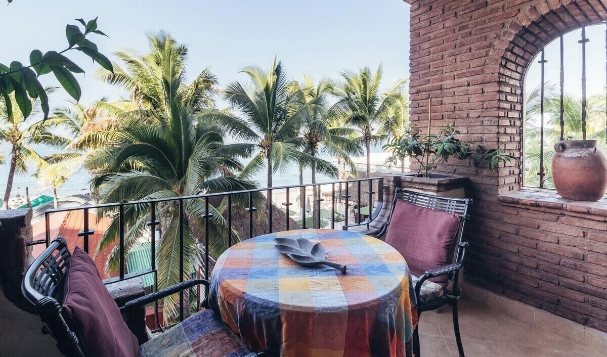 A balcony with a round table, chairs, and a view of palm trees and the sea.