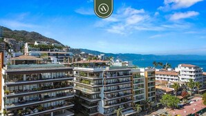 A multi-story hotel with balconies, located near a coastal area with a view of the sea and hills. - Elegant Studio in Romantic Zone - Rooftop Pool (Puerto Vallarta)